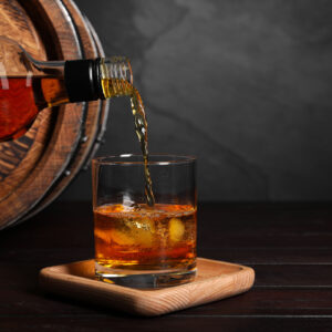 Pouring whiskey from bottle into glass with ice cubes near wooden barrel