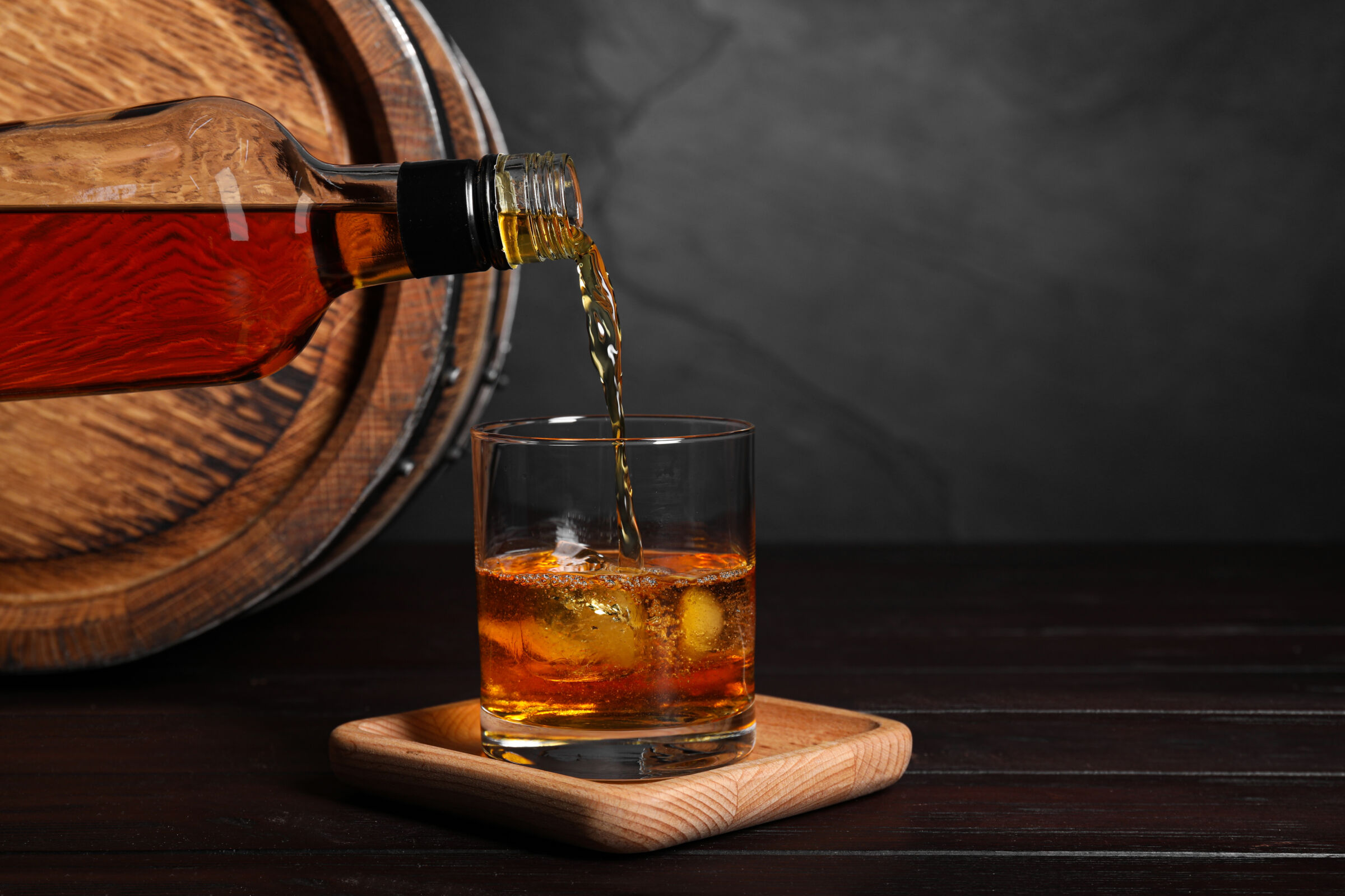 Pouring whiskey from bottle into glass with ice cubes near wooden barrel